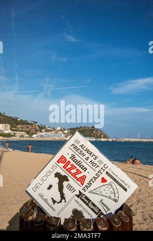 Pizza-Box zu Hause im Vordergrund mit dem Hintergrund des Strandes von Blanes mit Menschen, die sich dort unter einem wunderschönen blauen Sommerhimmel entspannen. Stockfoto