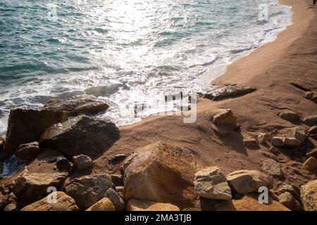 Wellen brechen gegen Steine im feinen Sand der Küste am Strand des Dorfes Blanes in Katalonien mit Reflexion der Sonne. Stockfoto