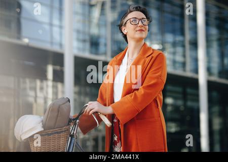 Eine nachdenkliche Geschäftsfrau, die ihr Fahrrad während ihres Morgenaufenthalts in der Stadt schiebt. Eine reife Geschäftsfrau, die aufmerksam wegsieht, während sie auf sie zugeht Stockfoto