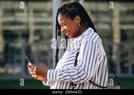 Lächelnde Geschäftsfrau, die auf dem Weg zur Arbeit in der Stadt ein Smartphone verwendet. Seitenansicht einer glücklichen jungen Geschäftsfrau, die auf eine SMS auf h antwortet Stockfoto