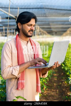 Vertikale Aufnahme von Happy Young Farmer beschäftigt sich mit der Arbeit an einem Laptop im Gewächshaus – Konzept von moderner Landwirtschaft, Technologie und Entwicklung oder Wachstum. Stockfoto