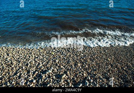 Ufer des Mittelmeers an einem der Steinstrände der Stadt White Coast Altea, in Alicante beobachten Sie, wie die Welle auf den Steinen endet. Stockfoto
