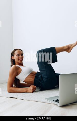 Eine Frau geht auf dem Boden Sport treiben, mit einem Online-Trainer über einen Laptop. Sport in Abriegelung Stockfoto