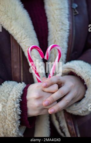 Nicht-binäre Person, die zwei weihnachtliche Zuckerstangen umklammert und ein Herz für den Valentinstag an einem kalten Wintertag mit einem Pullover macht. Stockfoto