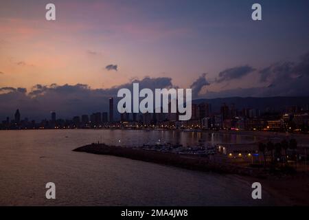 Sonnenuntergang mit violetten Farben am Himmel mit dunklen Wolken im Hintergrund über der Beleuchtung der Stadt Benidorm mit ihren Hotels. Stockfoto