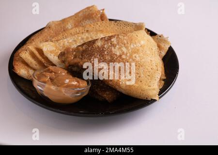 Foto von Pfannkuchen, die auf einem Teller liegen, und abgekochte Kondensmilch, die daneben auf weißem Hintergrund steht Stockfoto