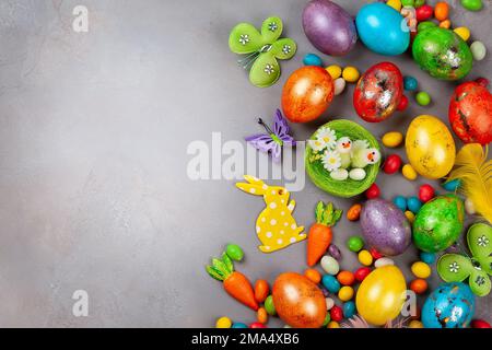 Farbenfrohe Ostereier, Schokoladen-Ostersüße, Häschen und Nest mit Küken auf grauem Hintergrund. Draufsicht mit Kopierbereich. Festliches Osterkonzept. Stockfoto