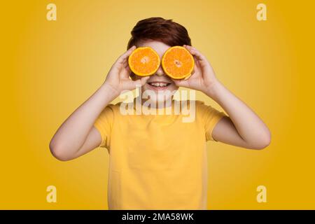 Ein rothaariger, lächelnder Junge, der vor seinen Augen die Hälften einer saftigen Orange auf gelbem Hintergrund hält Stockfoto