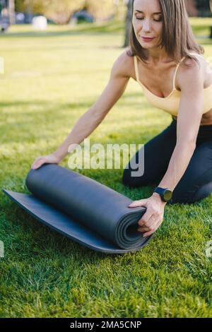 Junge Frau sitzt auf dem Gras und rollt Yoga-Matte und beendet das Outdoor-Workout am Morgen im Park. Ein glücklicher Lebensstil. Fitness-Frau. Stockfoto
