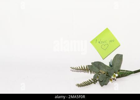 Eine Notiz, in der ich dich liebe, wurde von einem Liebhaber geschrieben, auf einem kleinen Blatt Grünpapier mit einem kleinen Blumenstrauß daneben. Stockfoto