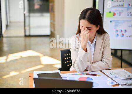Gestresste und depressive asiatische Millennials-Geschäftsfrau oder Buchhalterin, die sich Sorgen um ihre geschäftlichen Aufgaben macht und Kopfschmerzen oder Augenschmerzen hat. Stockfoto