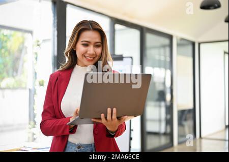 Professionelle und charmante asiatische Geschäftsfrau der Millennials steht in ihrem Büro mit einem tragbaren Laptop. Stockfoto