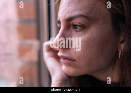 Nahaufnahme einer depressiven Frau, die aus dem Fenster schaut, während eine Träne ihre Wange runterfällt. Depression und psychisches Gesundheitskonzept Stockfoto