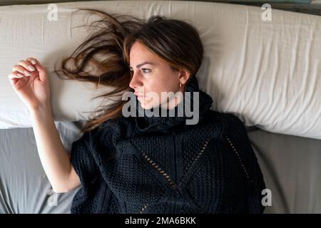 Blick von oben auf eine junge brünette Frau in einem schwarzen Kleid, die traurig aussieht und auf dem Bett liegt. Depression und psychisches Gesundheitskonzept Stockfoto