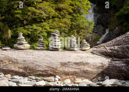 Horizontale Aufnahme von vielen Steinpyramiden auf einem alten Baumstamm im Wald und im Hintergrund des Wasserfalls. Ufer des Haast River. Brüllende Bi Stockfoto