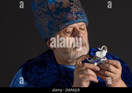 Nettes, unauffälliges Porträt eines bärtigen Seniorenmannes in blauer orientalischer Kleidung, der eine Elefantenfigur aus Porzellan mitnimmt und es sich ansieht Stockfoto