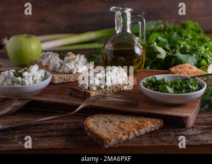 Geröstetes Sauerteigbrot mit Hüttenkäsesalat auf einem Holztisch mit Kräutern und Olivenölkaraffe Stockfoto