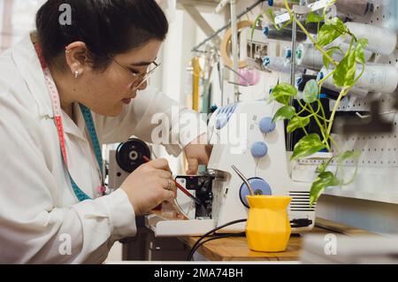 Junge argentinische Näherin in ihrem Nähatelier, sitzt, reinigt ihre Nähmaschine und bereitet sie auf die Arbeit vor. Stockfoto