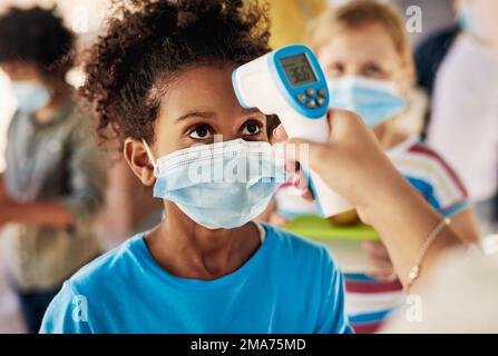 Deine Temperatur ist in Ordnung. Ein junges Mädchen, das eine Maske trägt und seine Temperatur überprüfen lässt, bevor es in der Pause zum Spielen rausgeht. Stockfoto