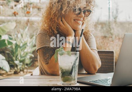 Eine moderne digitale Mitarbeiterin genießt es, im Sommer mit einem Cocktail mohito im Café zu sitzen und an einem Laptop und einer Online-Internetverbindung zu arbeiten. Di Stockfoto