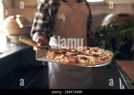 Direkt aus dem Ofen und in deinen Bauch. Ein unbekannter Mann, der allein steht und eine frisch zubereitete Pizza in seinem Restaurant hält. Stockfoto