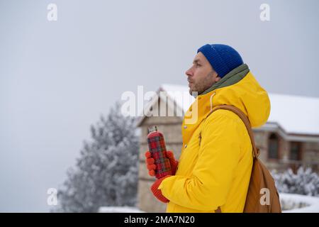 Ein nachdenklicher Mann geht in der Nähe von zu Hause mit einem heißen Getränk in einer Wärmflasche und denkt an den eisigen Wintertag nach. Stockfoto