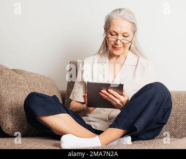 Ältere Frau mit grauen Haaren und Brille, die mit gekreuzten Beinen auf einer Couch sitzt und ein digitales Tablet benutzt Stockfoto