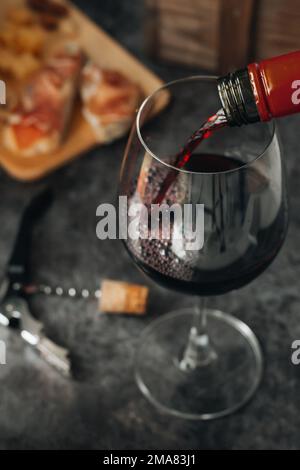 Rotwein aus der Flasche in Glas. Ein Glas Rotwein liegt auf dem Tisch. Prosciutto mit Käse, Rotwein, Baguette, Nüssen. Rotwein mit Tradition Stockfoto