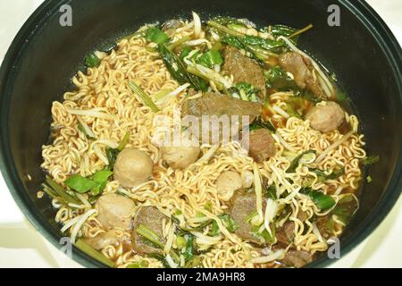 Gekochte Instantnudeln, geschmortes Rindfleisch und Fleischbällchen in schwarzer Kräutersuppe auf einem elektrischen Kochtopf Stockfoto