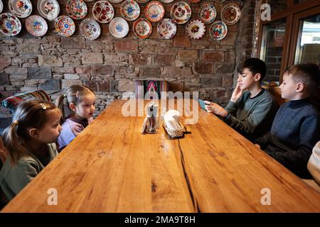 Vier Kinder sitzen in einem authentischen ukrainischen Restaurant und schauen auf Telefone. Stockfoto