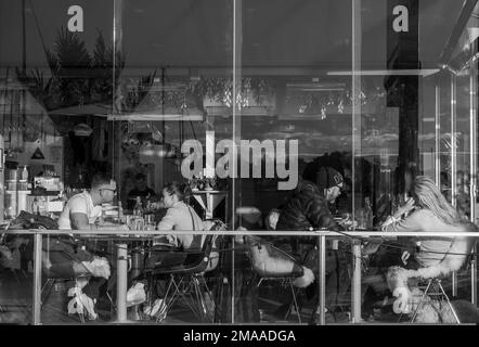 Junge Paare sitzen an Tischen in der Kongen Marina Beach Bar & Restaurant, die an einem sonnigen Tag durch eine Glasfassade mit Reflexionen gesehen werden. Oslo, Norwegen. Stockfoto