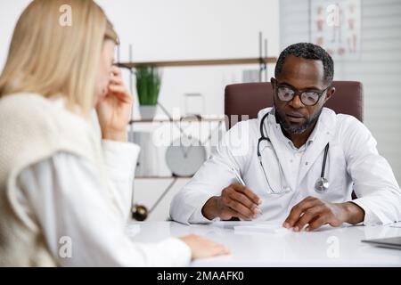 Afroamerikanischer Arzt, empfängt einen Patienten, hört sich Beschwerden an, stellt Diagnosen an. Schreibt Daten. Arzttermin. Stockfoto