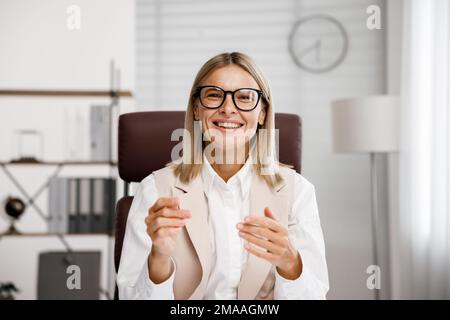 Lächelnde Geschäftsfrau mittleren Alters, die sich Webcam-Videokonferenzen ansieht, in einem virtuellen Chat-Meeting anruft und von dort aus einen Webinar-Kurs abhält Stockfoto