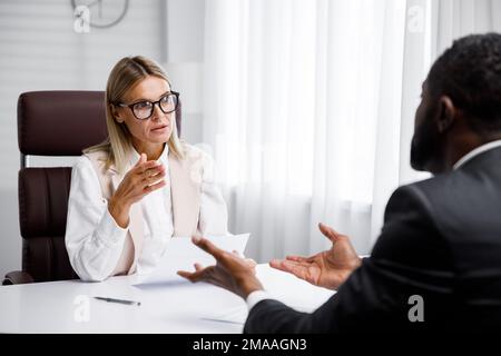 Einstellung, Vorstellungsgespräch. Blonde Geschäftsfrau mittleren Alters, die ein Interview in einem hellen Büro führt, ein afroamerikanischer Mann. Er sieht sich seinen Lebenslauf an und Stockfoto