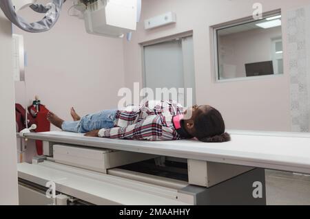 Ein afroamerikanisches Mädchen ist eine Patientin, die auf der Oberfläche eines Lehrlings für eine Röntgenuntersuchung liegt. Stockfoto