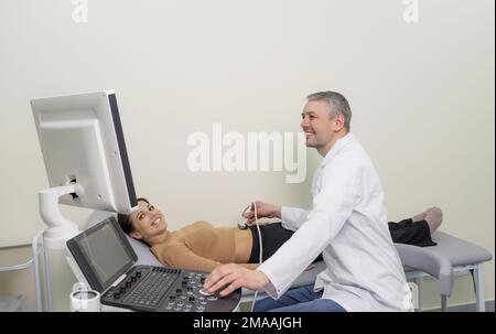 Ein männlicher Arzt bewegt einen Ultraschallsensor auf dem Magen einer schwangeren Frau, während er in einem Krankenhaus auf einen Bildschirm schaut Stockfoto