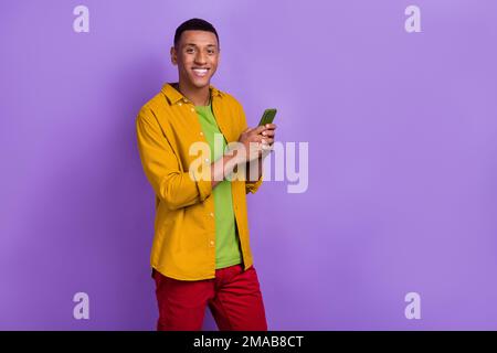 Foto eines lustigen positiven Mannes trägt ein gelbes Hemd und tippt Apfel samsung modernes Gerät leerer Raum isolierter violetter Hintergrund Stockfoto