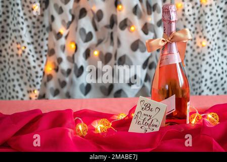Rosa Champagner mit einer romantischen Note auf einem Tisch mit einem satinroten Tuch gefüllt mit Glühbirnen auf schwarzem und weißem herzförmigem Hintergrund. Stockfoto