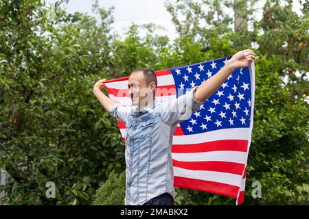 Positiv attraktiver amerikanischer junger Mann schlang sich in die Flagge Amerikas und schaut in die Kamera, im Freien Stockfoto