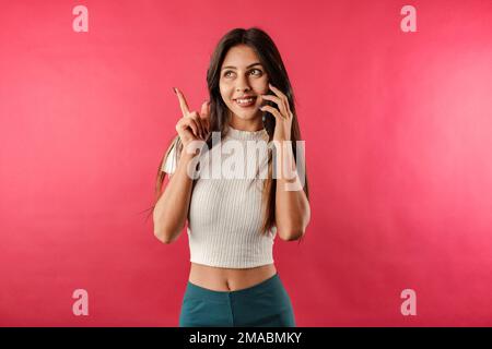 Frau lächelt selbstbewusst, isoliert über roten Hintergrund, spricht am Telefon, schaut auf den Kopierbereich und zeigt mit dem Zeigefinger. Anzeige. Pointi Stockfoto