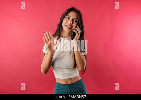 Junge, dunkelhaarige Frau mit gerippter, isolierter Frucht auf rotem Hintergrund, die am Telefon spricht und mit der Hand winkt. Ich schaue weg. Stockfoto