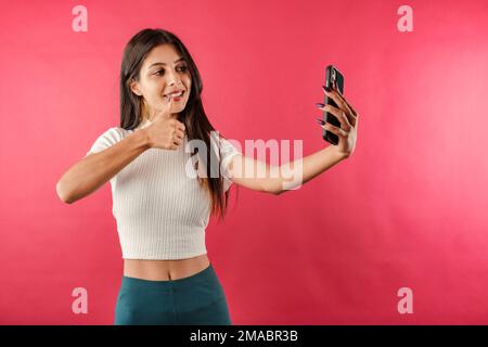 Süße, dunkelhaarige Frau, die ein lässiges Oberteil trägt, isoliert auf rotem Hintergrund, Selfie und Gesten wie Daumen hoch zum Smartphone macht, mit lebendiger Stimme kommuniziert Stockfoto