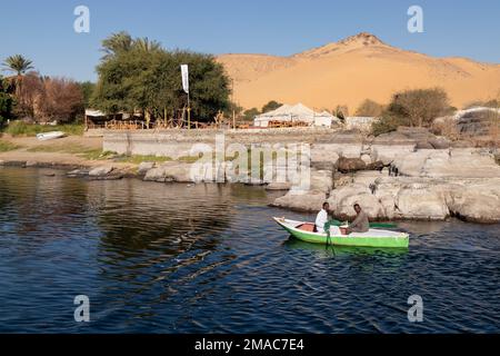 ASSUAN, ÄGYPTEN - 29. Dez. 2022. Zwei nubische Männer paddeln in einem hölzernen Boot nahe der Küste in Ägypten mit sandiger Berglandschaft im Hintergrund Stockfoto