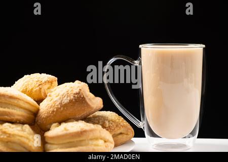 Tee mit Milch in einem Glasbecher mit doppeltem Boden und Kuchen auf schwarzem Hintergrund Stockfoto