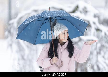 Eine glückliche Frau hält einen Schirm am kalten, verschneiten Tag. Schneeflocken fallen auf ihre Hand. Stockfoto