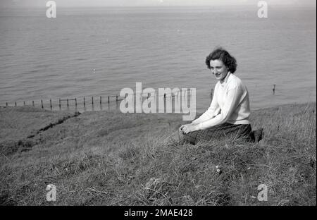 1950er, historisch, eine attraktive junge Dame, Mitte zwanzig Jahre alt, trägt ein Oberteil aus Baumwolle mit Rollkragen und einen Rock mit Schottenmuster, sitzt für ein Foto auf einem grasbedeckten Hügel mit Blick auf das Meer, England, Großbritannien. Stockfoto