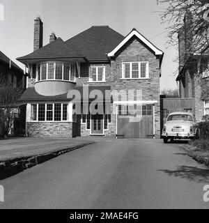 1960er, historisch, Vorderansicht eines großen Vorstadthauses, mit interner Garage, mit Holztüren, England, Großbritannien. Ein Woseley Auto der Ära parkt in der Auffahrt vor der zweiten Garage, die an der Seite des Hauses gebaut wurde und an das benachbarte Grundstück angeschlossen ist. Stockfoto