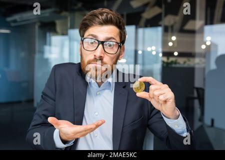 Nahaufnahme. Verwirrter junger Geschäftsmann hat eine Goldmünze, Kryptowährung, Bitmünze in der Hand. Er schaut in die Kamera, zeigt auf eine Münze, zeigt mit der Hand, stellt eine Frage. Stockfoto