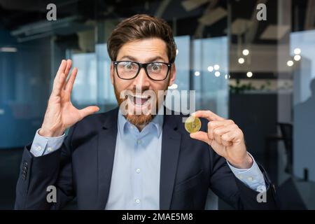 Konzept, Nahaufnahme. Ein junger Geschäftsmann hält eine Goldmünze, Kryptowährung in der Hand. Er schaut in die Kamera und schreit vor Glück Stockfoto