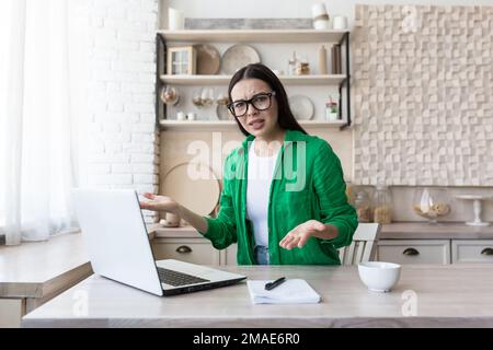 Wütende junge Frau, die zu Hause am Küchentisch vor dem Laptop sitzt, mit Notizbuch und Dokumenten. Er sieht verwirrt in die Kamera und spreizt seine Hände. Stockfoto